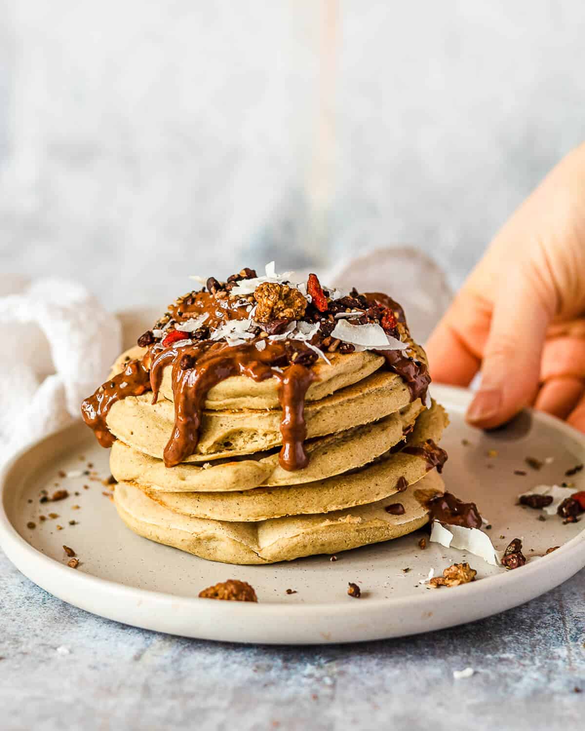A stack of 5 vegan buckwheat pancakes on a grey plate. On top of the pancakes there is some cacao hazelnut butter, coconut chips and various seeds. There is a hand holding the plate.