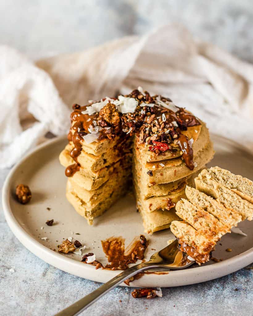 A stack of 5 vegan buckwheat pancakes on a grey plate. On top of the pancakes there is some cacao hazelnut butter, coconut chips and various seeds. A section of the pancake stack has been cut and is on a fork in front of the pancakes.