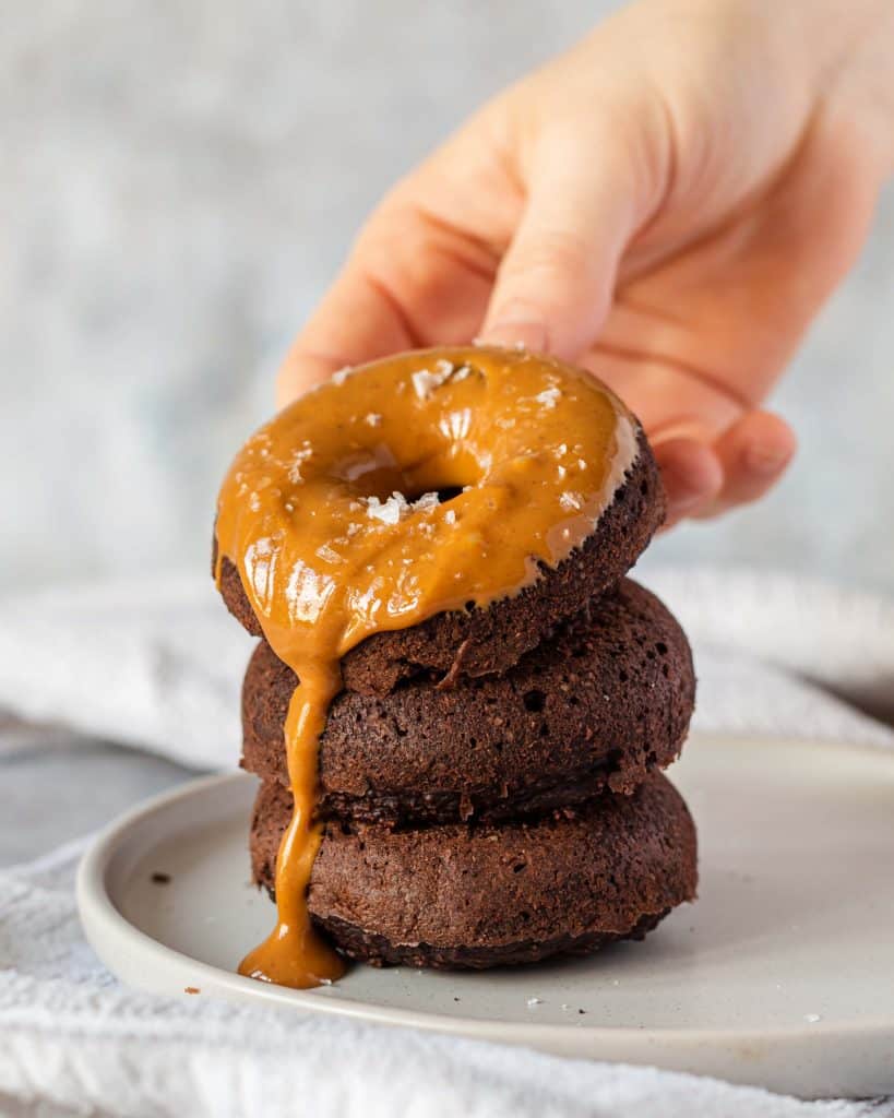 Three Chocolate Baked Protein Donuts stacked on top of each other on a grey plate, there is peanut butter spread on the top on and it's dripping down the side. There is a hand holding the top one.