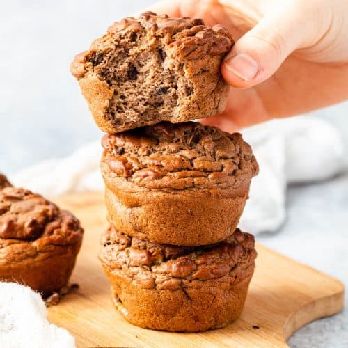 Four Banana Buckwheat Muffins on a bread board, three are stacked on top of each other. A bite has been taken from the top muffin and a hand is holding it. There is another muffin to the left.