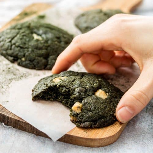 Three Spirulina Cookies on a wooden chopping board. A bite has been taken from the front cookie and there is a hand holding it.