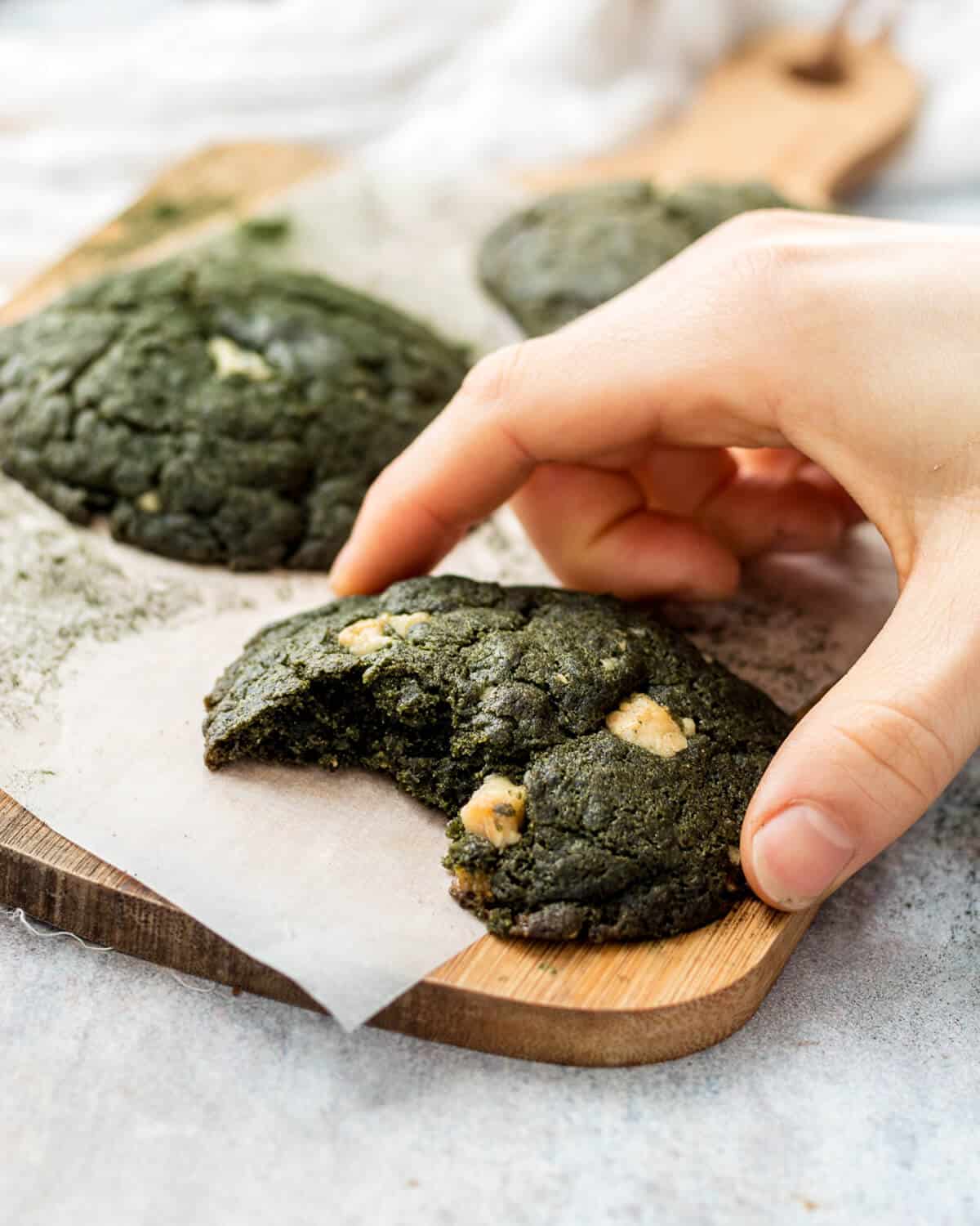 Three Spirulina Cookies on a wooden chopping board. A bite has been taken from the front cookie and there is a hand holding it.