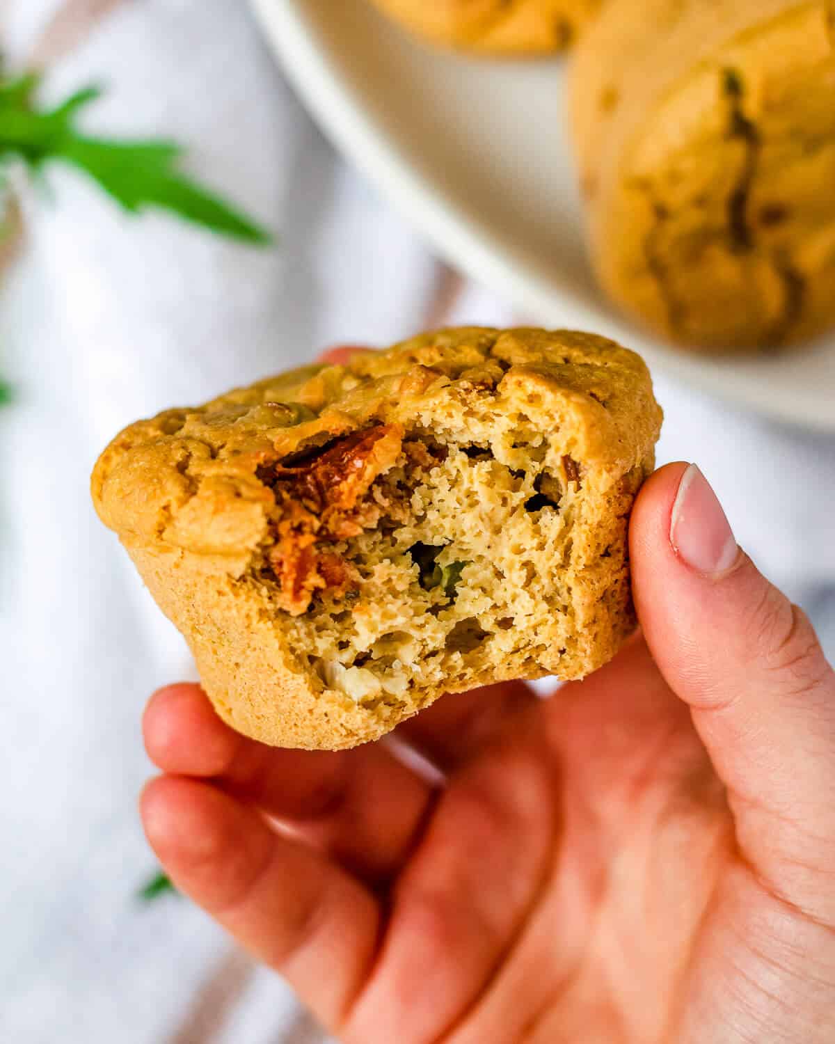 A hand holding a savoury chickpea flour muffin - a bite has been taken revealing the fluffy texture and a sun-dried tomato. There are more muffins on a plate in the background.