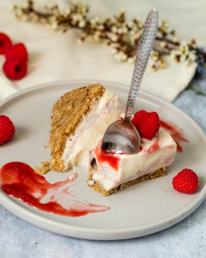 Two slices of vegan white chocolate cheesecake with raspberry coulis on a grey plate. There is a bite missing from one of the slices and there is a teaspoon standing upright in the slice. In the background there are some flowers as decoration.