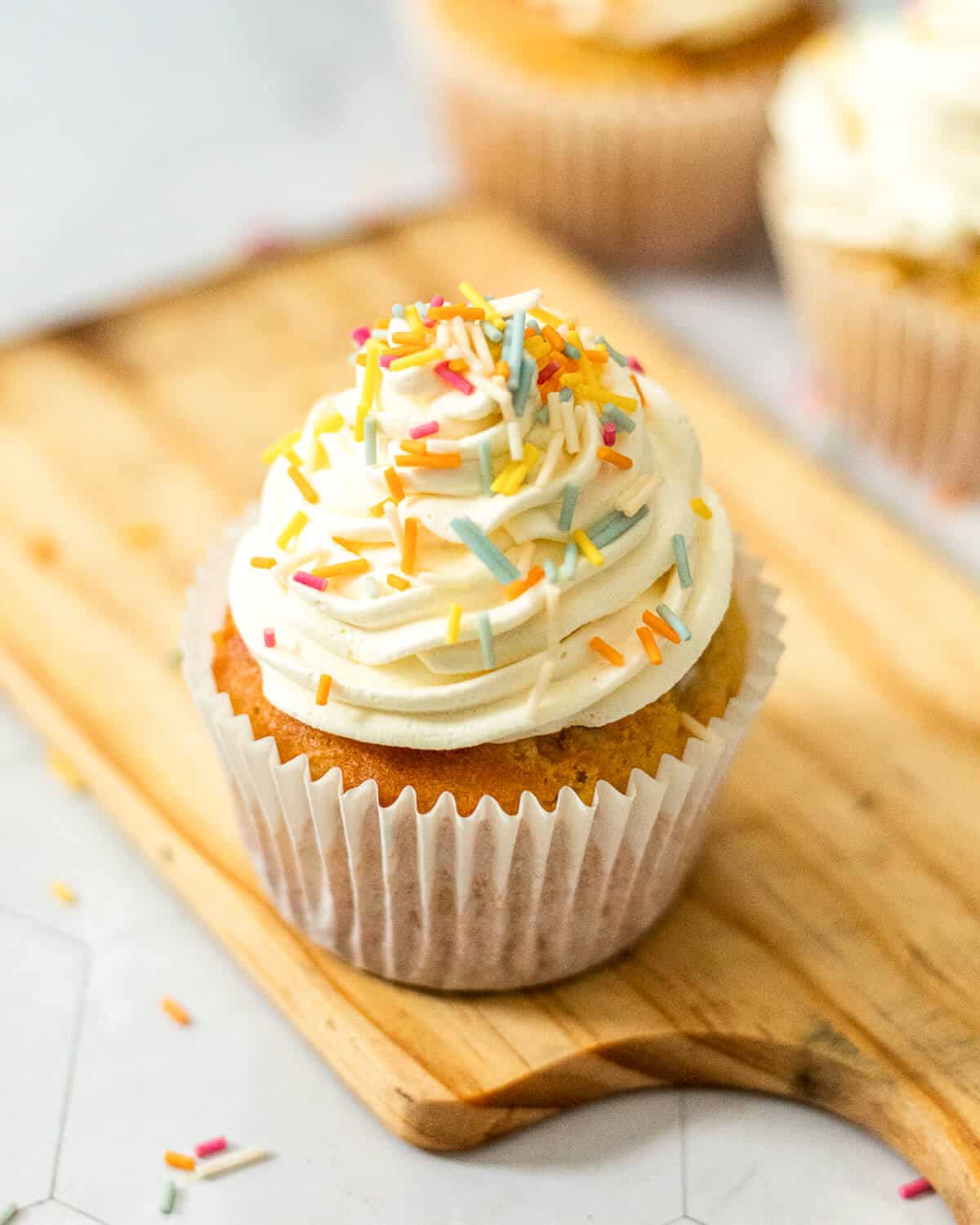 A vanilla cupcake with funfetti sprinkles on a small wooden board. There are two more cupcakes in the background.