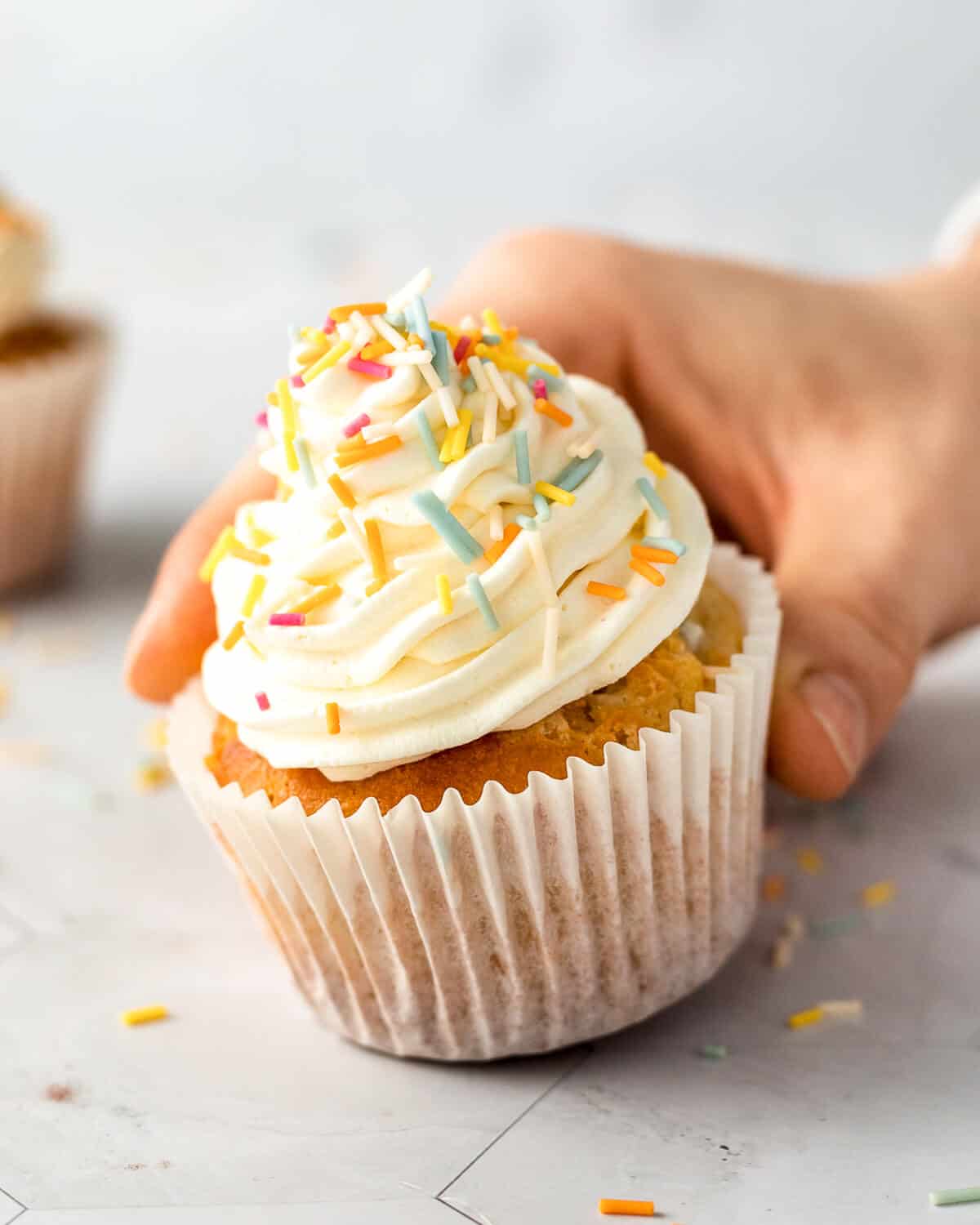 A hand holding an easy vanilla cupcake with sprinkles, there is another cupcake in the background.