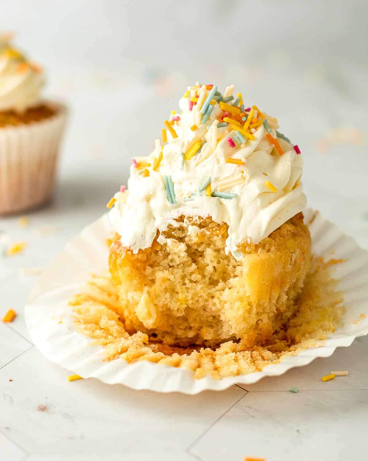 A vanilla birthday cupcake with a bite taken in a cupcake liner. There is another cupcake in the background.