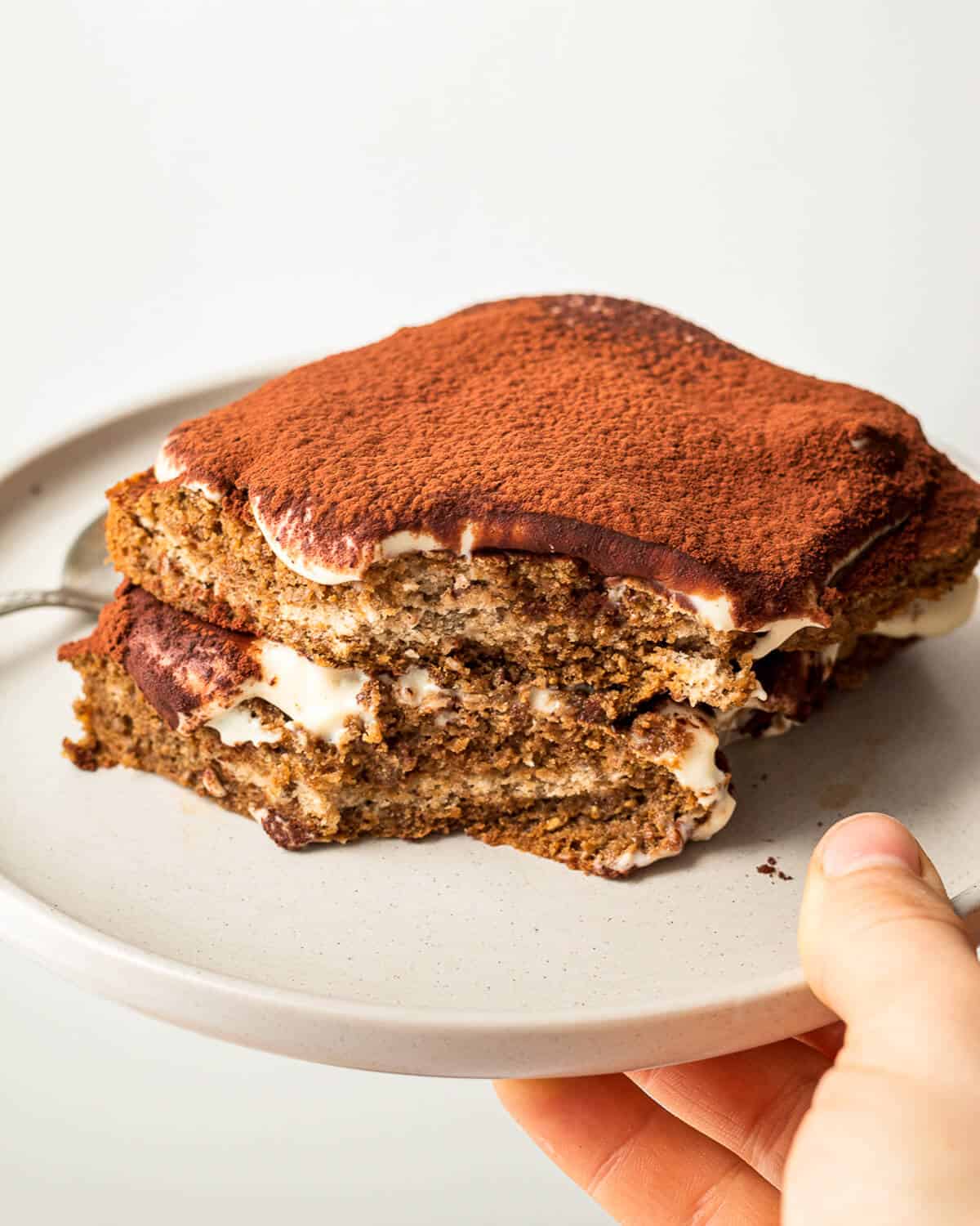 A hand holding a grey plate with a single serve tiramisu with a bite missing.