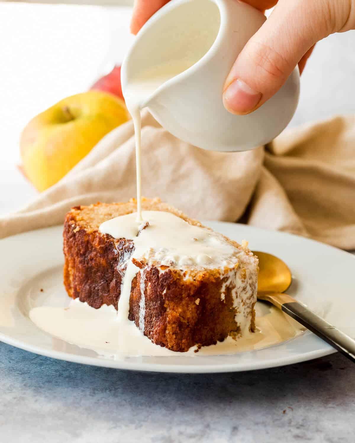 A slice of Apple and Cinnamon Cake on a white plate, someone is pouring cream on the cake from a white jug. There is an appl ein the background and a teaspoon on the plate.