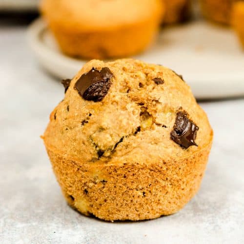 A banana muffin with chocolate chips, there are more muffins on a plate in the background.