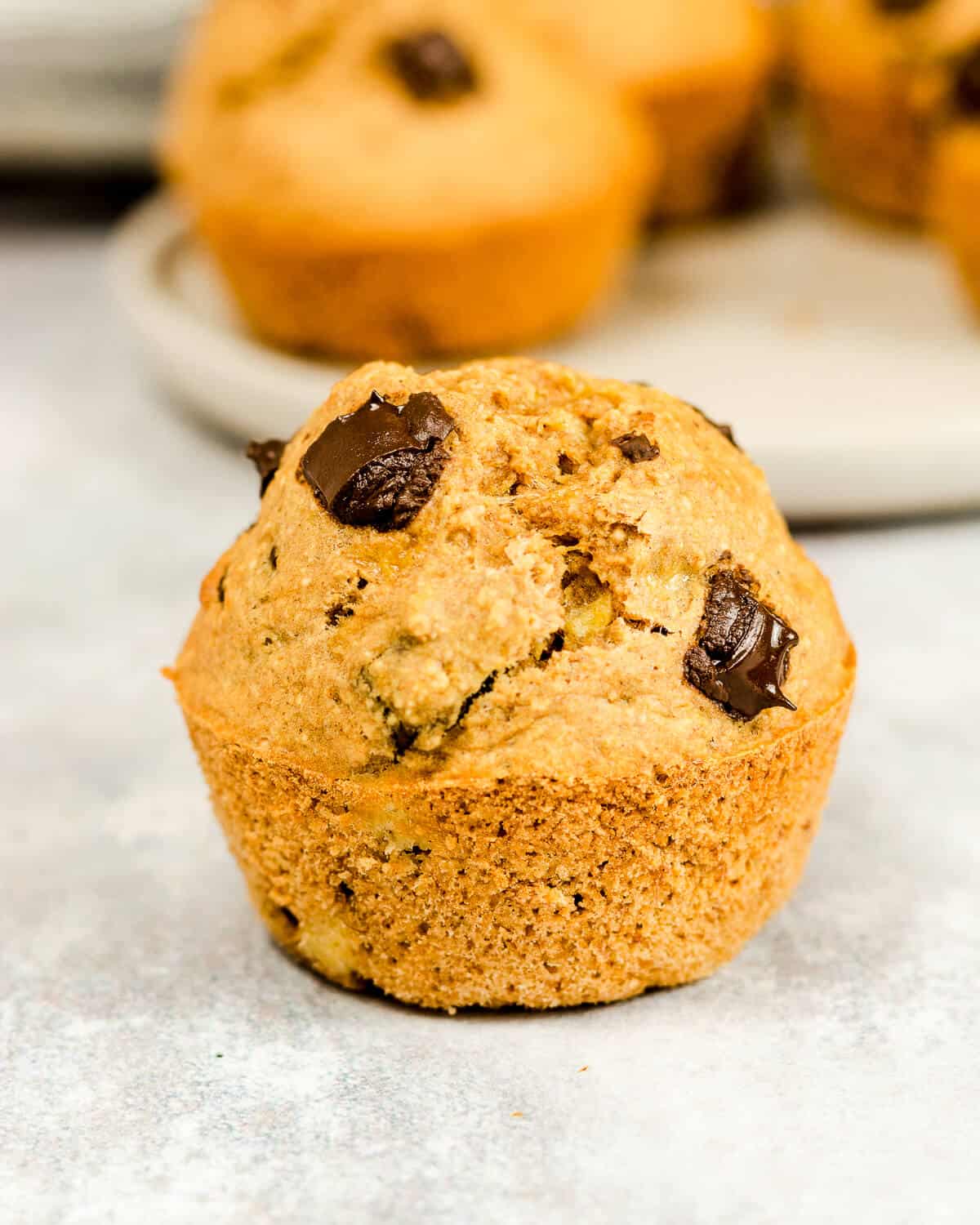 A banana muffin with chocolate chips, there are more muffins on a plate in the background.