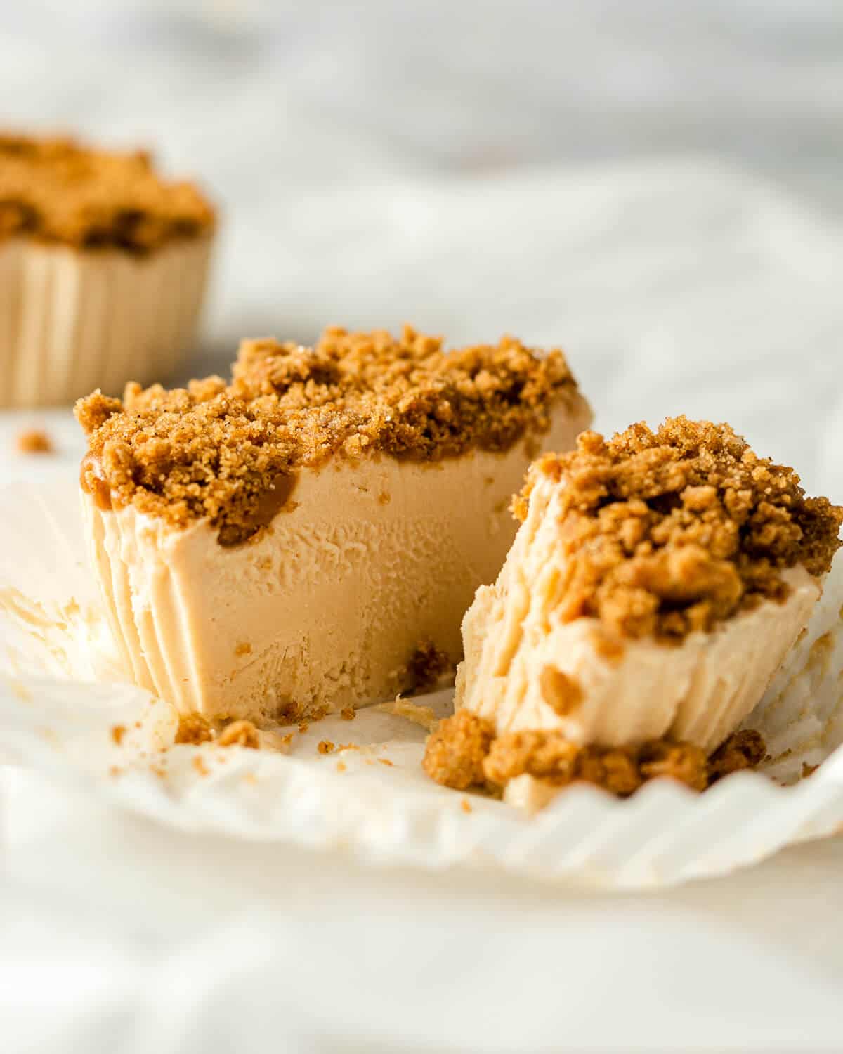 A mini biscoff cheesecake in a cupcake liner is cut in half and a bite is missing from one of the halves. There is another cheesecake in the background.