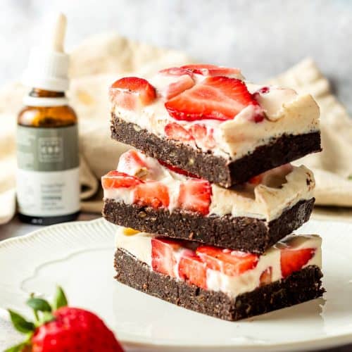Three cheesecake brownies with strawberries stacked on top of each other on a white plate. There is a strawberry as a prop and a bottle of CBD oil in the background.