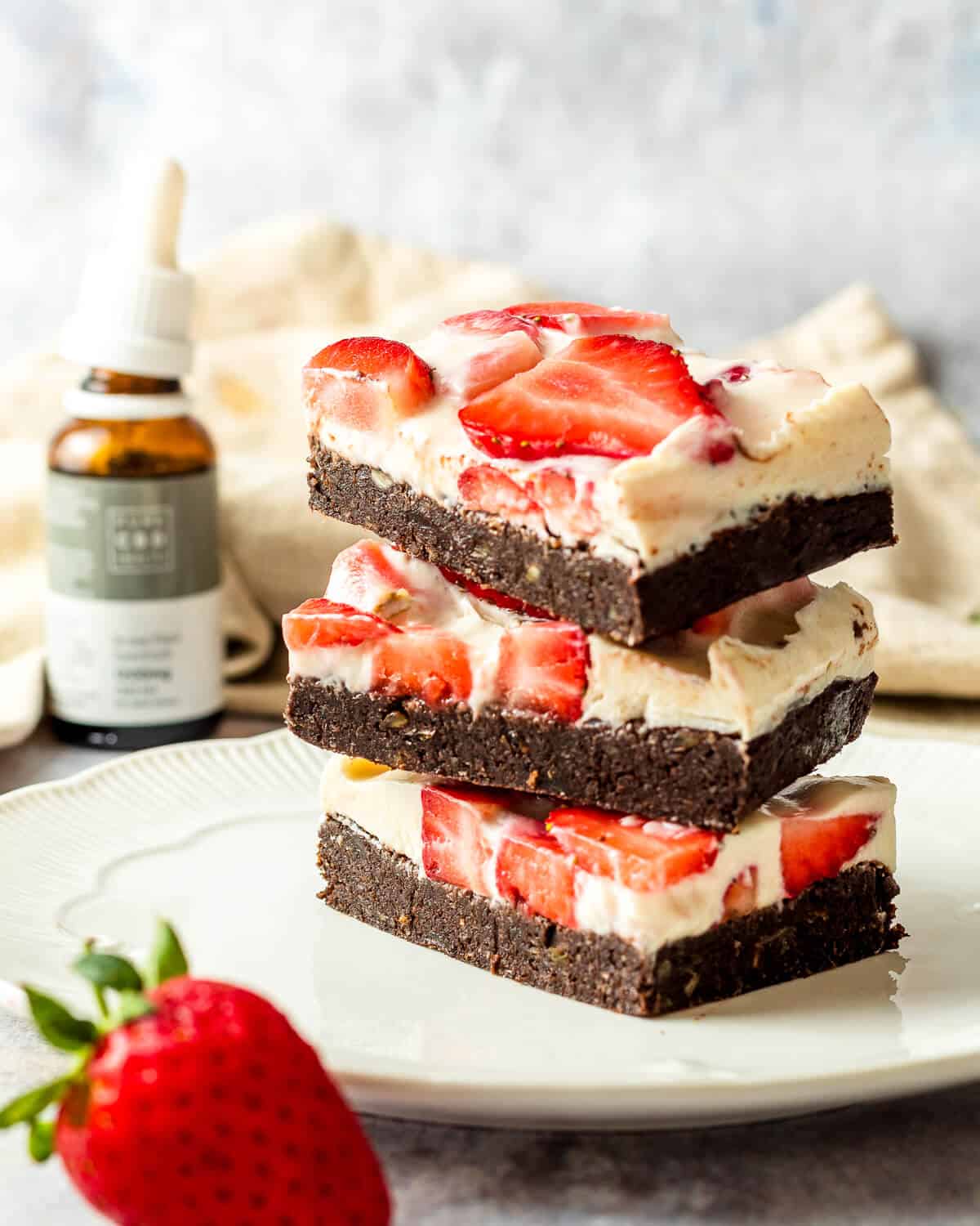 Three cheesecake brownies with strawberries stacked on top of each other on a white plate. There is a strawberry as a prop and a bottle of CBD oil in the background.