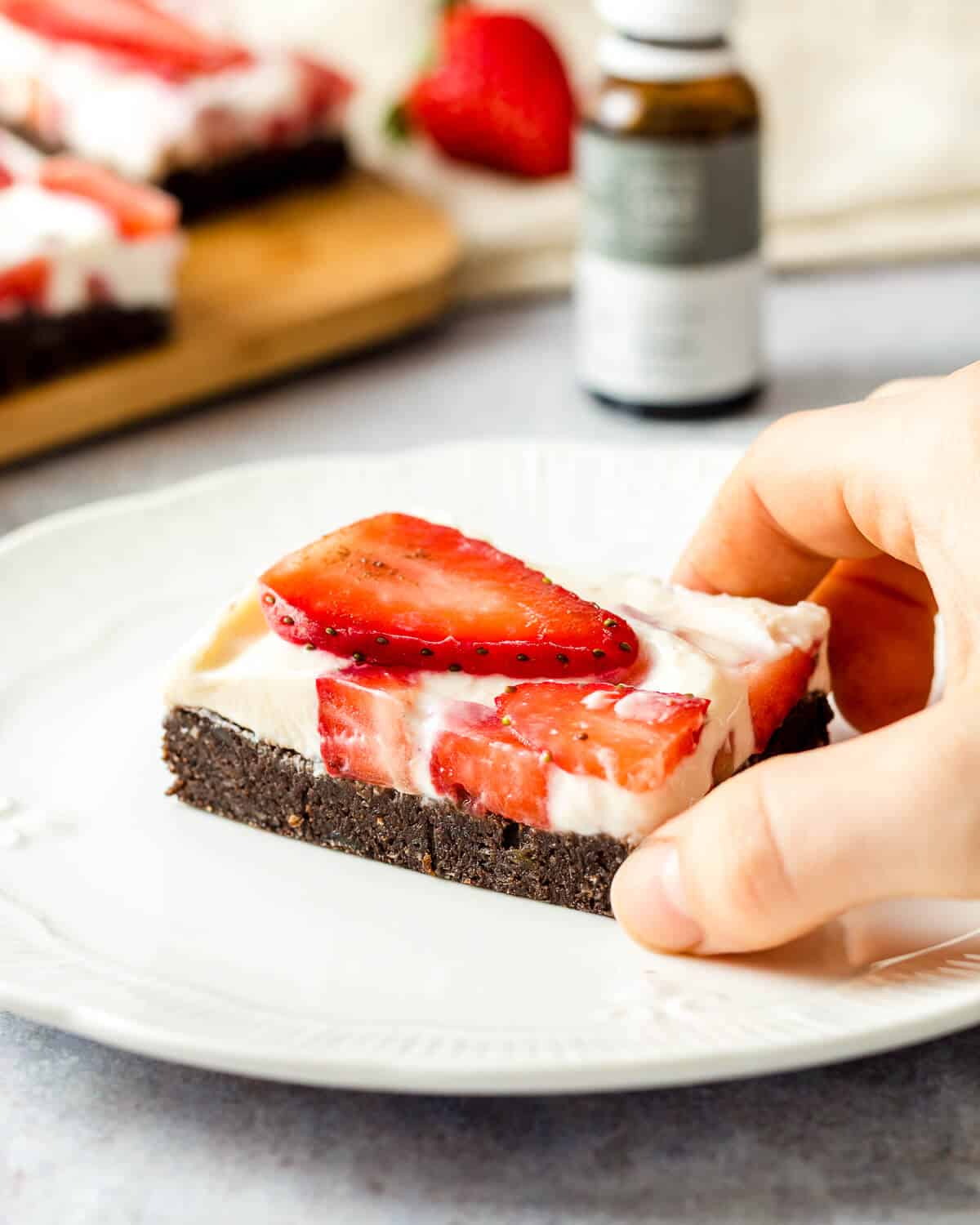 A hand is holding a no bake cheesecake brownie with strawberries on a white plate. There are more brownies in the background as well as a bottle of CBD oil.