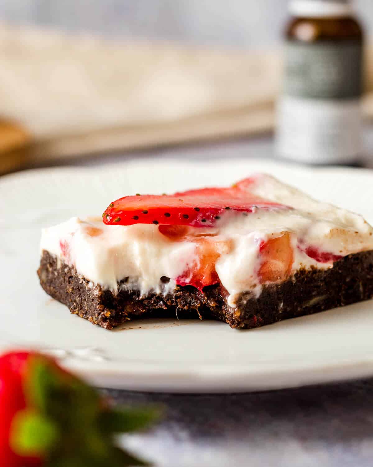 A cheesecake brownie with strawberries on a white plate. A bite has been taken. There is a bottle of CBD oil in the background.