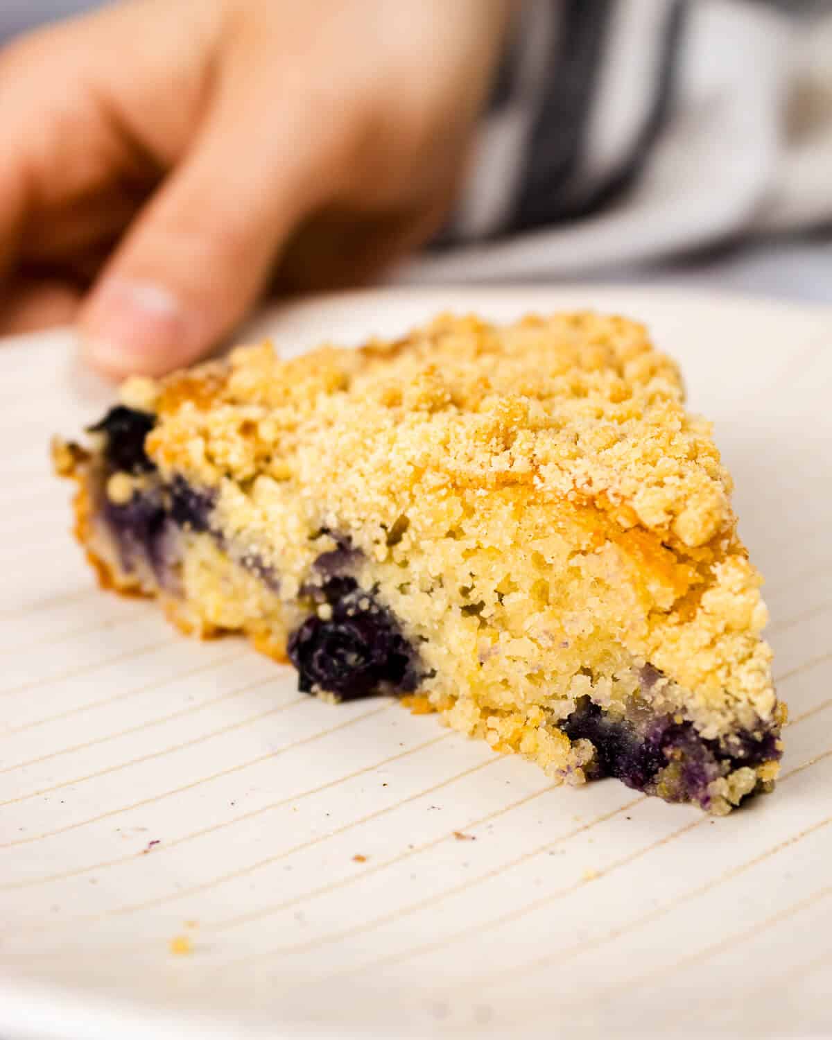 A slice of blueberry crumble cake of a white plate, a hand is holding the plate.