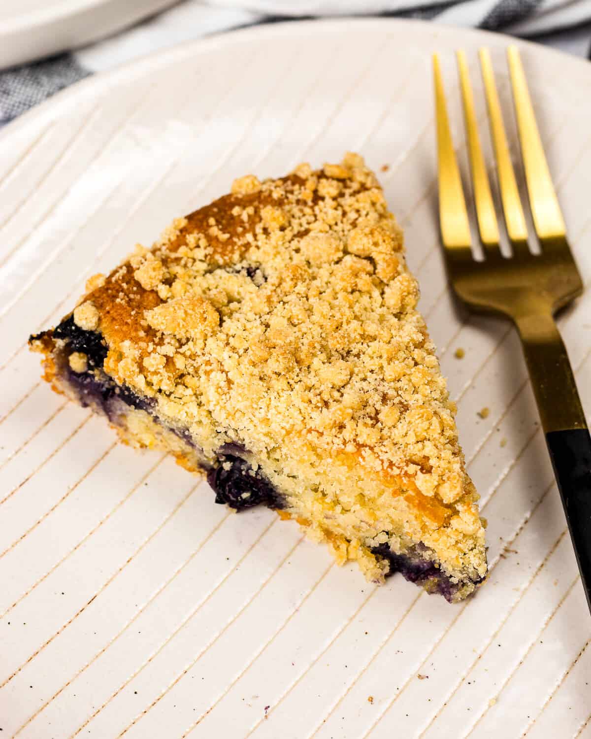 A slice of blueberry crumble cake on a white plate with a fork on the right.