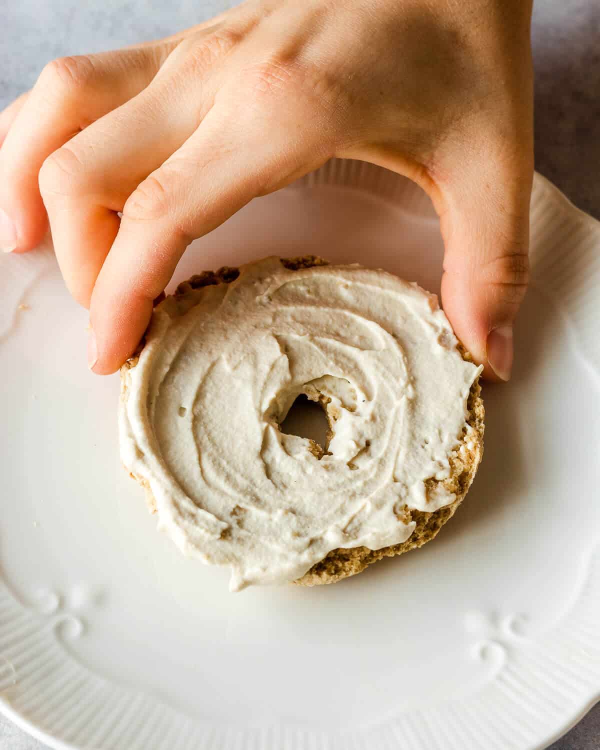 A hand is grabbing half a bagel with cream cheese on a white plate.