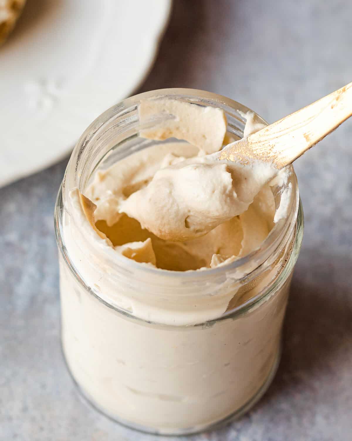 A jar of vegan cream cheese. There is a knife coming out of the jar with some cream cheese on it. There is a plate in the background.