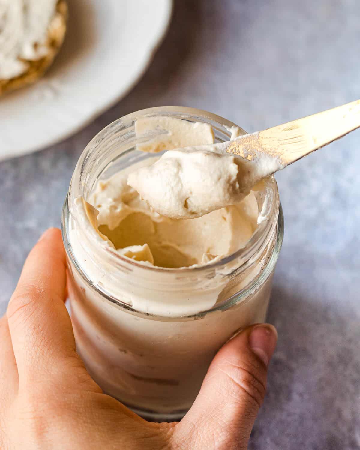 A jar of vegan cream cheese. Someone is holding the jar and a knife with some cream cheese on it. There is a plate in the background.