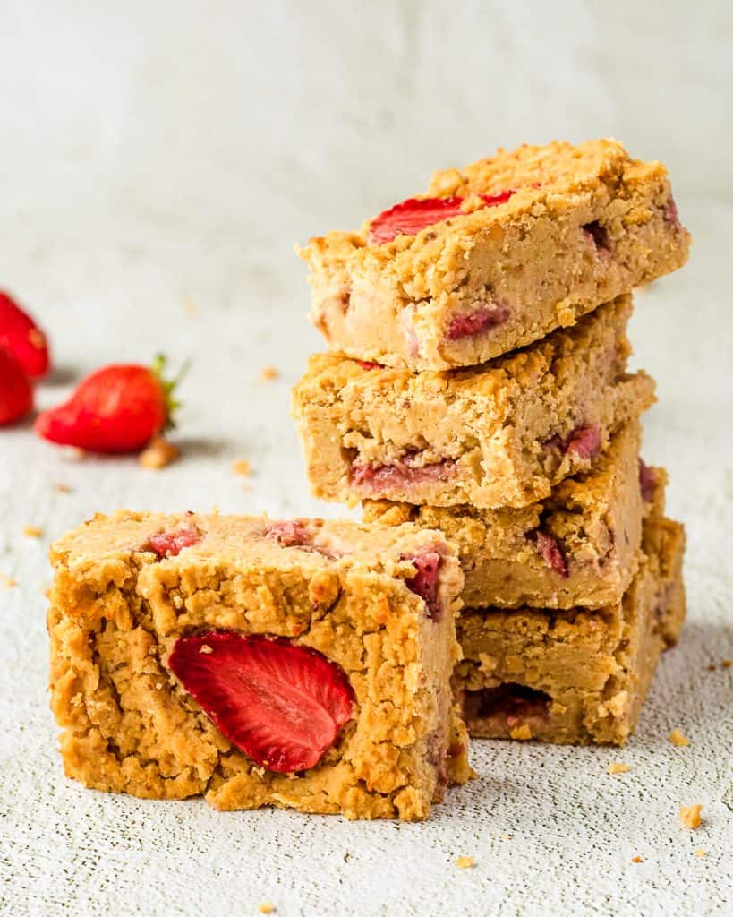 A photo of five chickpea blondies with strawberries. There are four are stacked on top of each other and another placed in front. There are strawberries in the background.