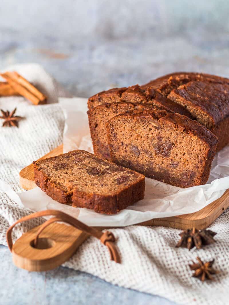 A Fruit Cake Loaf on a sheet of white baking parchment paper on a wood cutting board. There are some whole spices in the background.