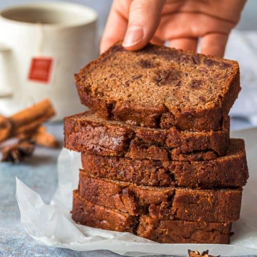 Five fruit cake loaf slices stacke on top of each other, there is a hand holding the top slice. In the background is a cup of tea and various whole spices.