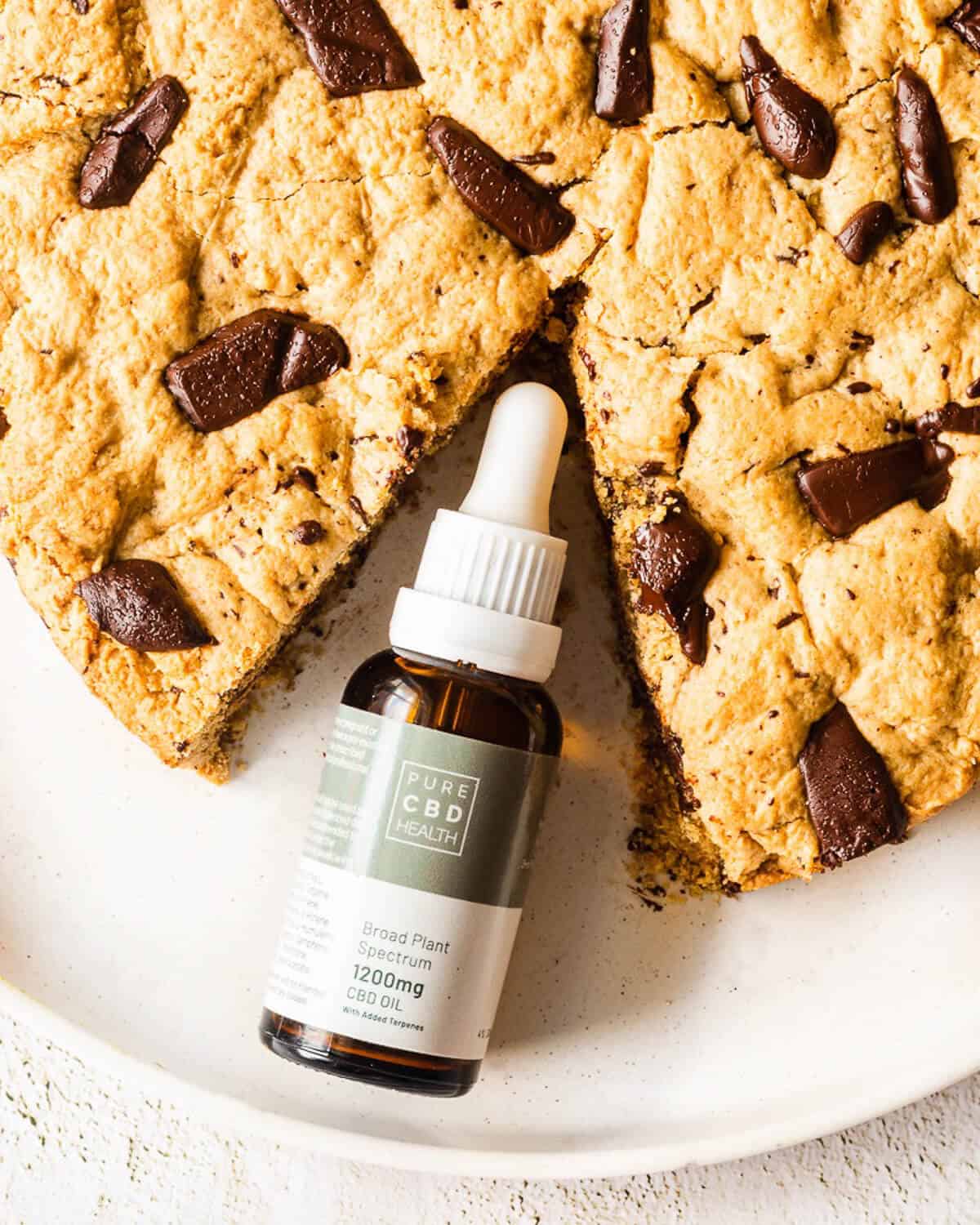 A Giant Chocolate Chip Cookie on a white plate, a slice is missing and in its place is a bottle of cbd oil.