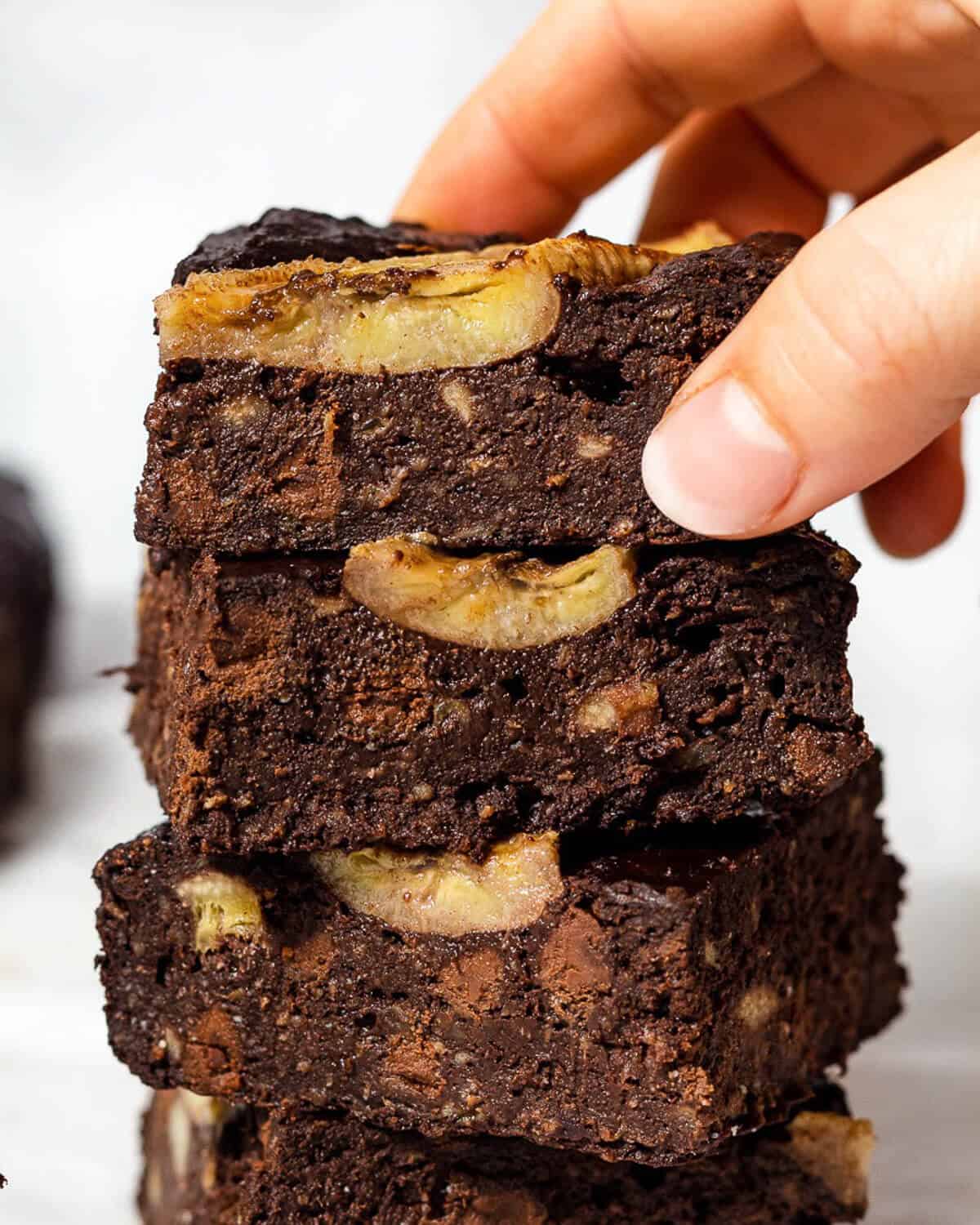 Four fudgy banana brownies with chocolate chips are stacked on top of each other, a hand is holding the brownie on top.
