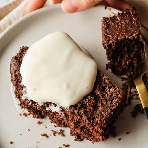 A hand holding a white plate with a slice of chocolate loaf cake. There is some vegan yoghurt on the cake and a bite has been taken from the cake which is on a fork to the right.