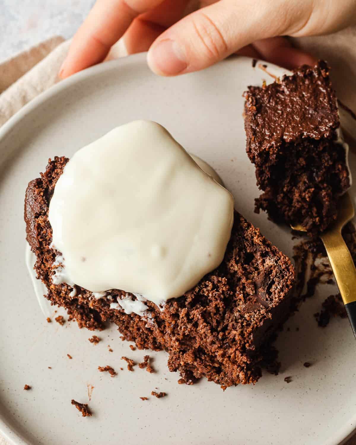 A hand holding a white plate with a slice of chocolate loaf cake. There is some vegan yoghurt on the cake and a bite has been taken from the cake which is on a fork to the right.