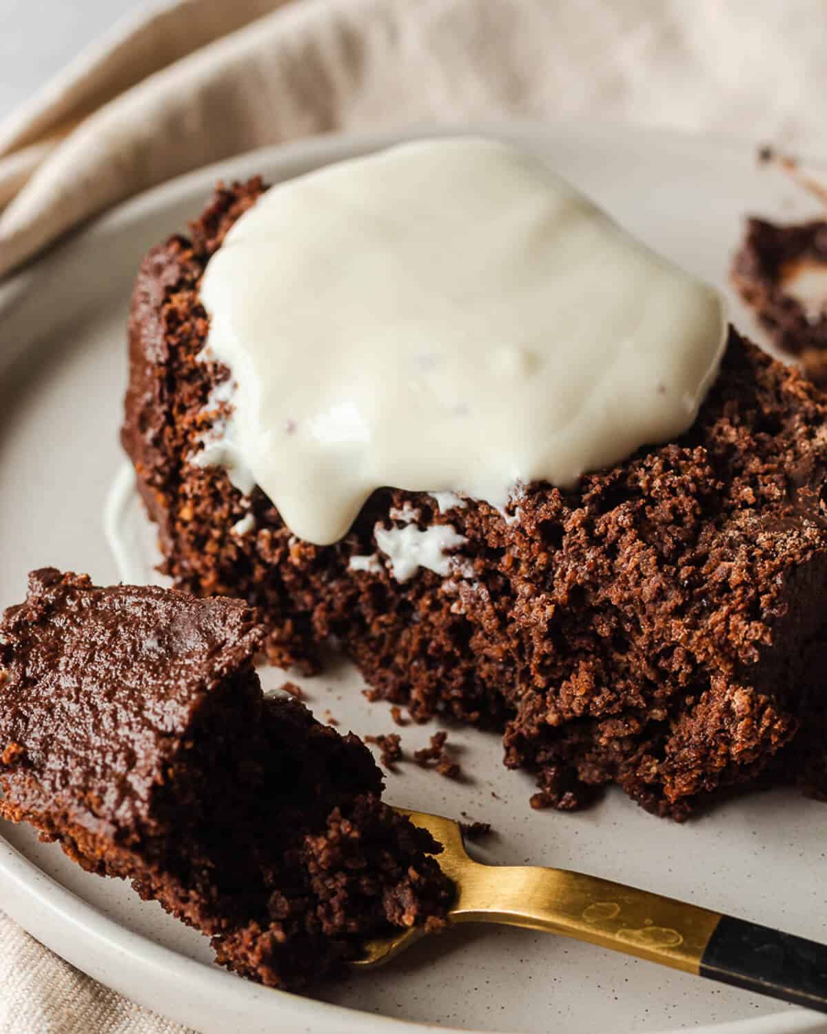 A hand holding a white plate with a slice of chocolate loaf cake. There is some vegan yoghurt on the cake and a bite has been taken from the cake which is on a fork in front of the cake.