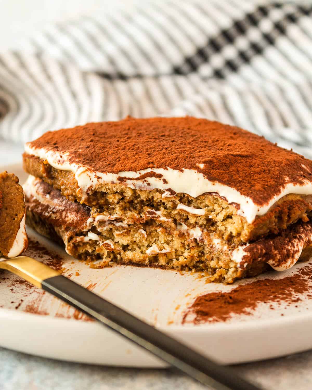 A single serving of tiramisu on a grey plate. There is a fork to the left of the tiramisu. A section of the tiramisu has been eaten.