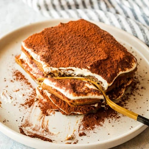 A single serving of tiramisu on a grey plate. There is a fork cutting a section of the tiramisu off.