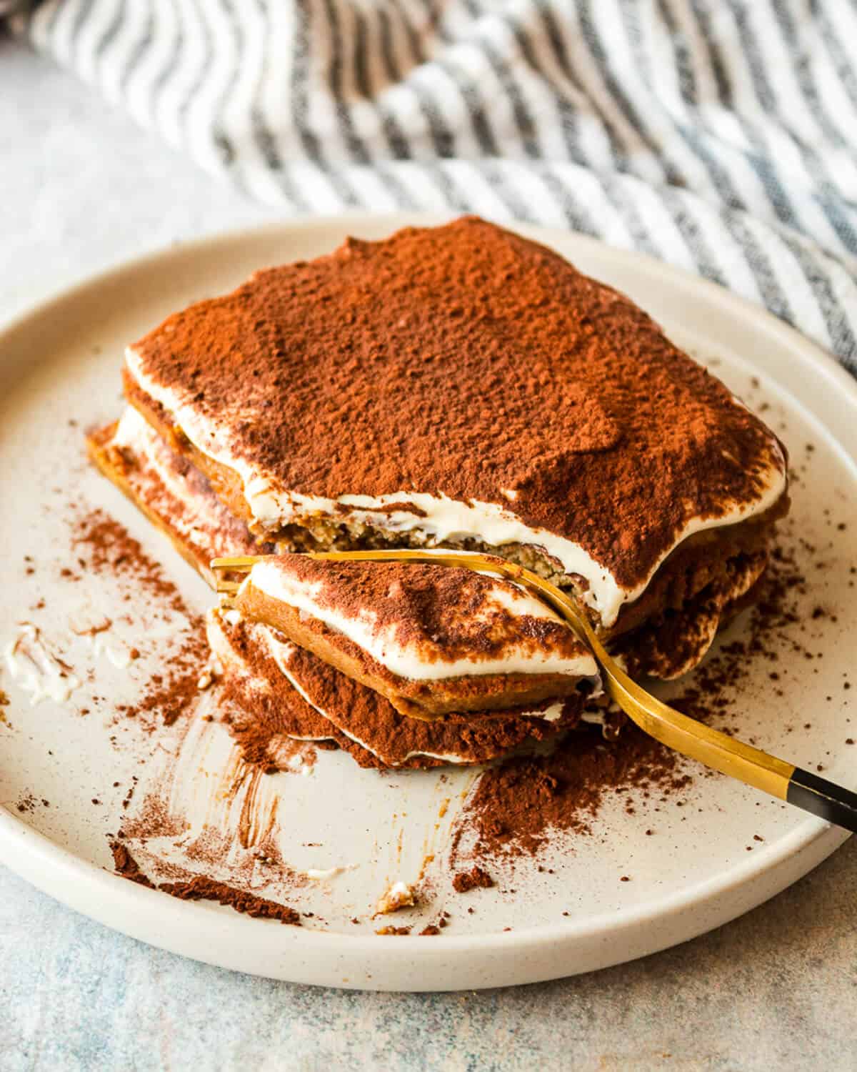 A single serving of tiramisu on a grey plate. There is a fork cutting a section of the tiramisu off.
