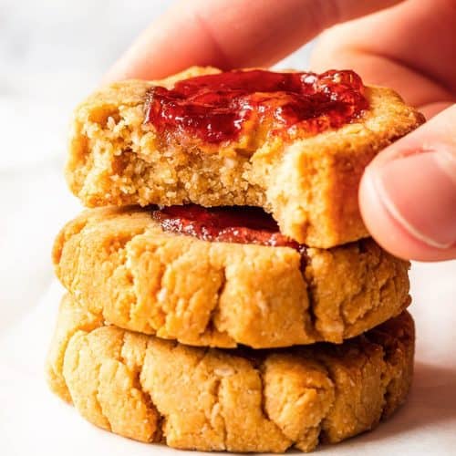 Three Jam Thumbprint Cookies stacked on top of each other, a bite has been taken from the top cookie revealing the soft texture. Someone is holding the top cookie.