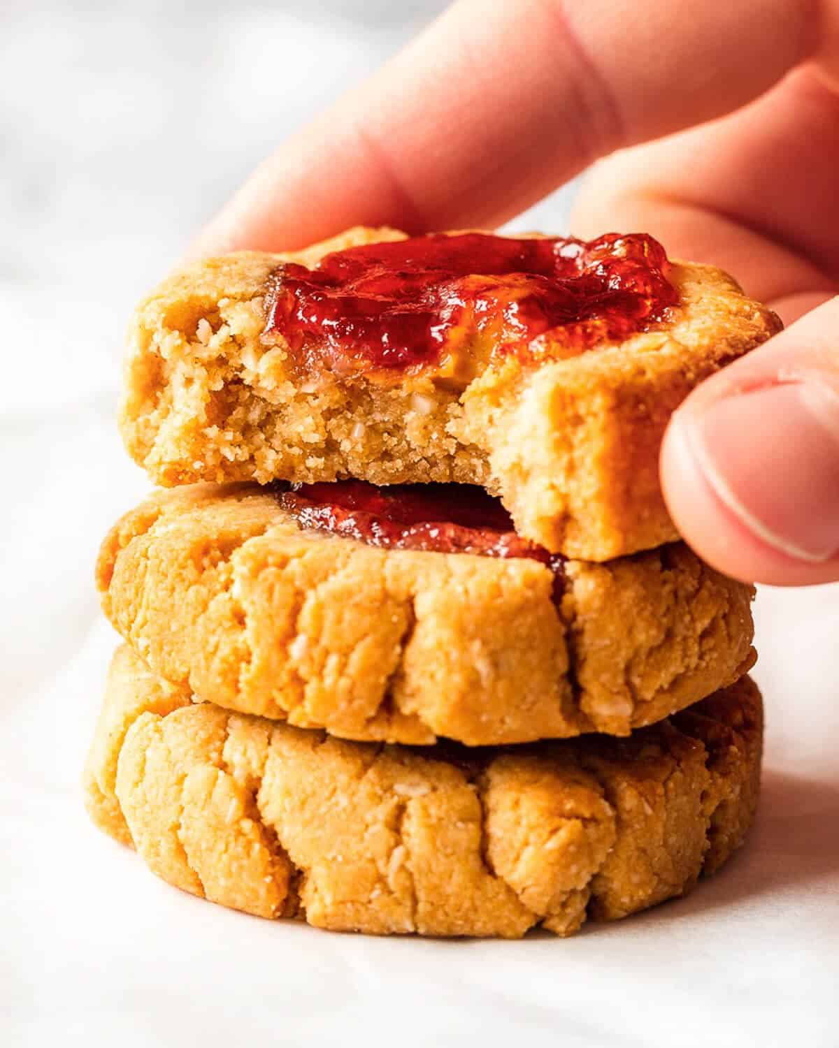 Three Jam Thumbprint Cookies stacked on top of each other, a bite has been taken from the top cookie revealing the soft texture. Someone is holding the top cookie.