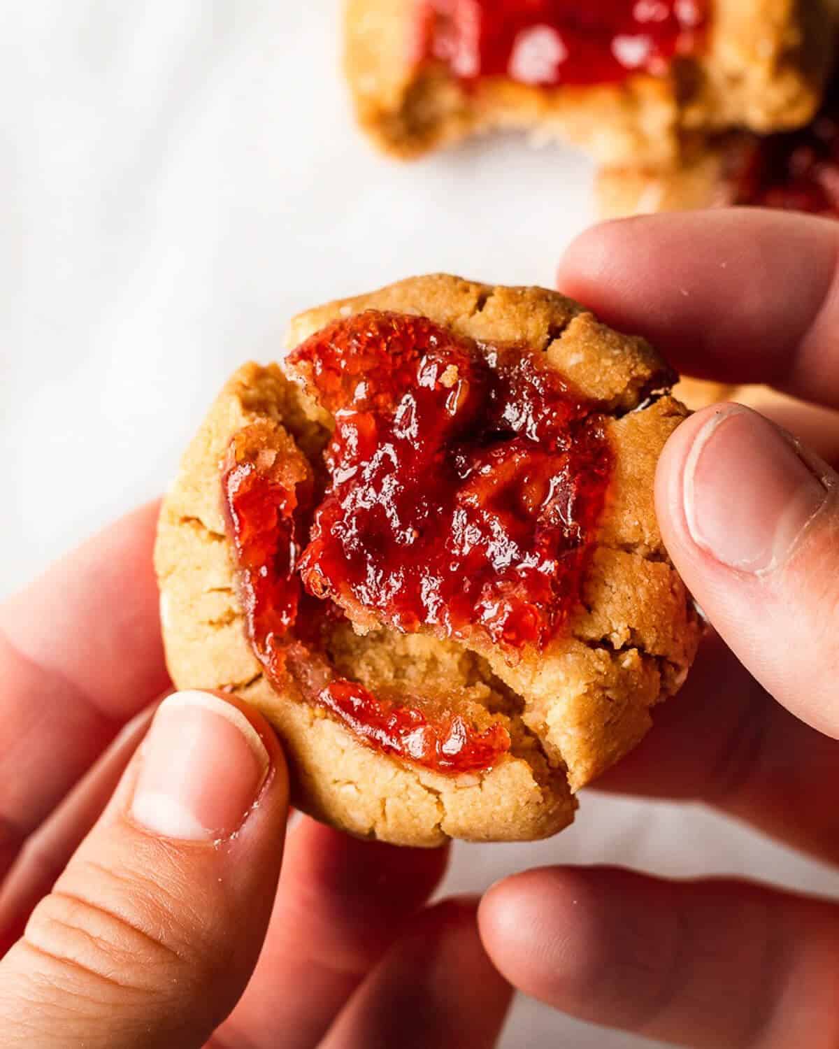 Two hands holding a jam thumbprint cookie, they're breaking it in half. There are a few more cookies in the background.