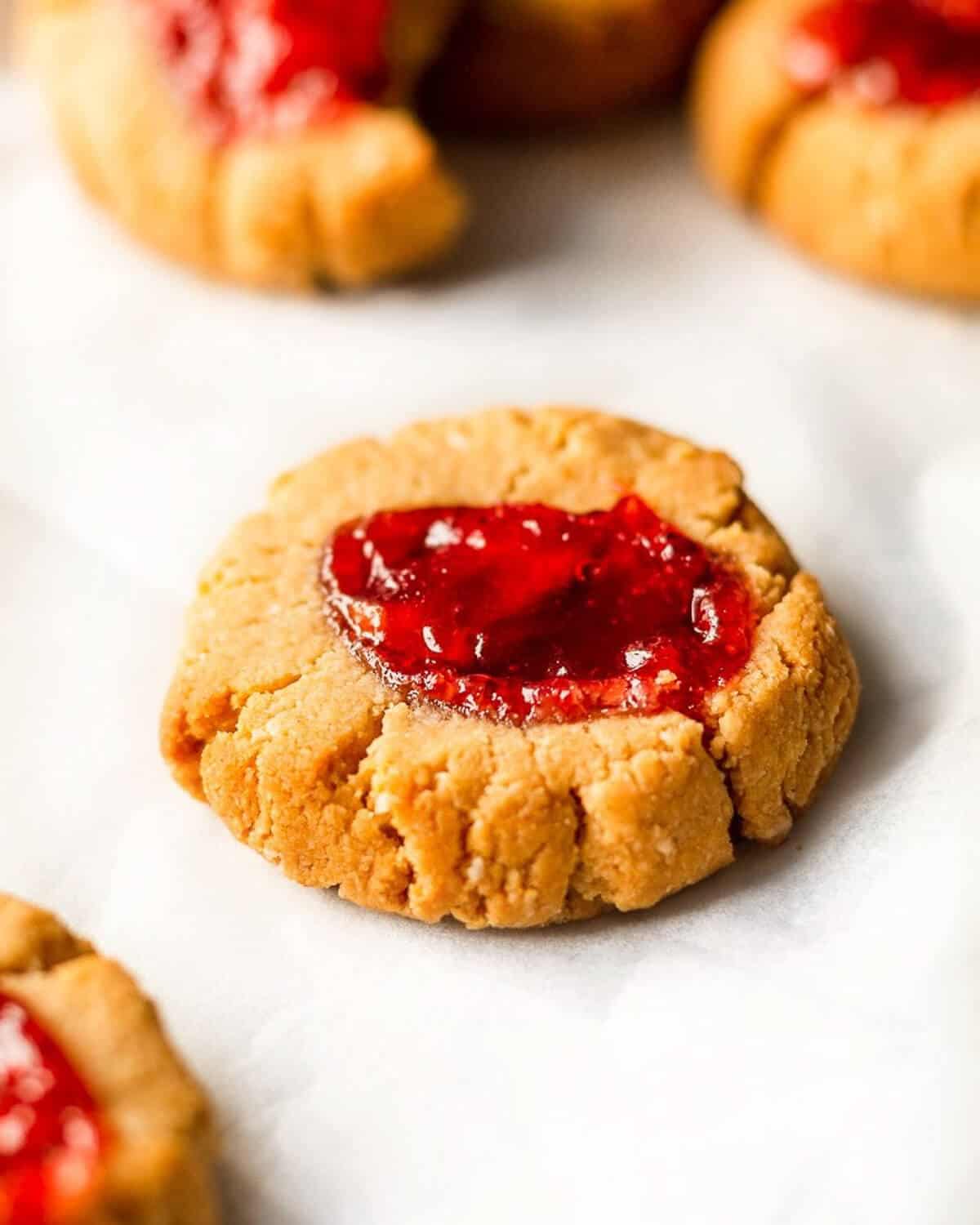 A PB & J Thumbprint Cookie is in focus in the middle of the photo, there are a few more cookies out of focus in the background.