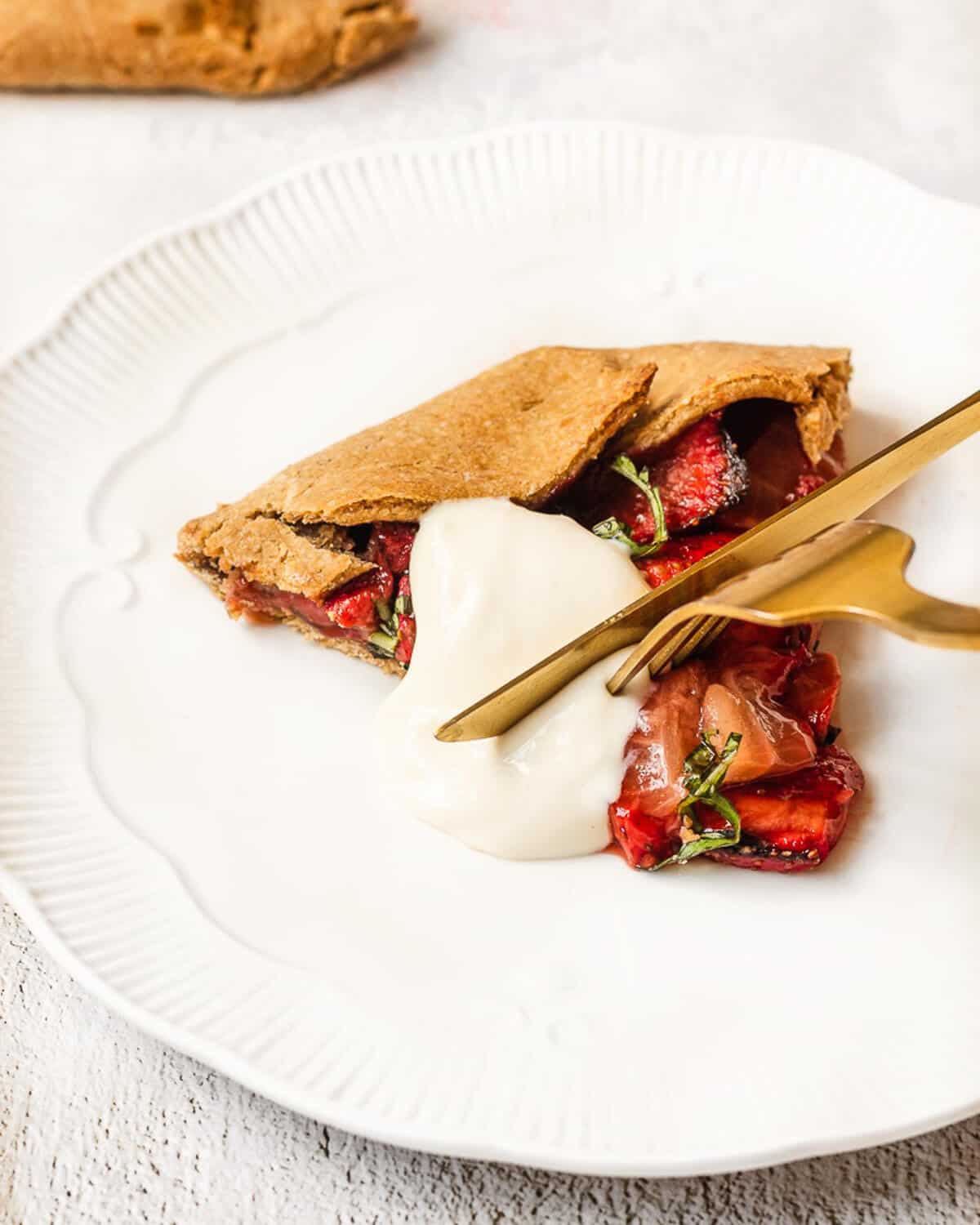 a knife and fork is cutting a bite of a slice of strawberry galette on a white plate. There is some vegan yoghurt on the galette.