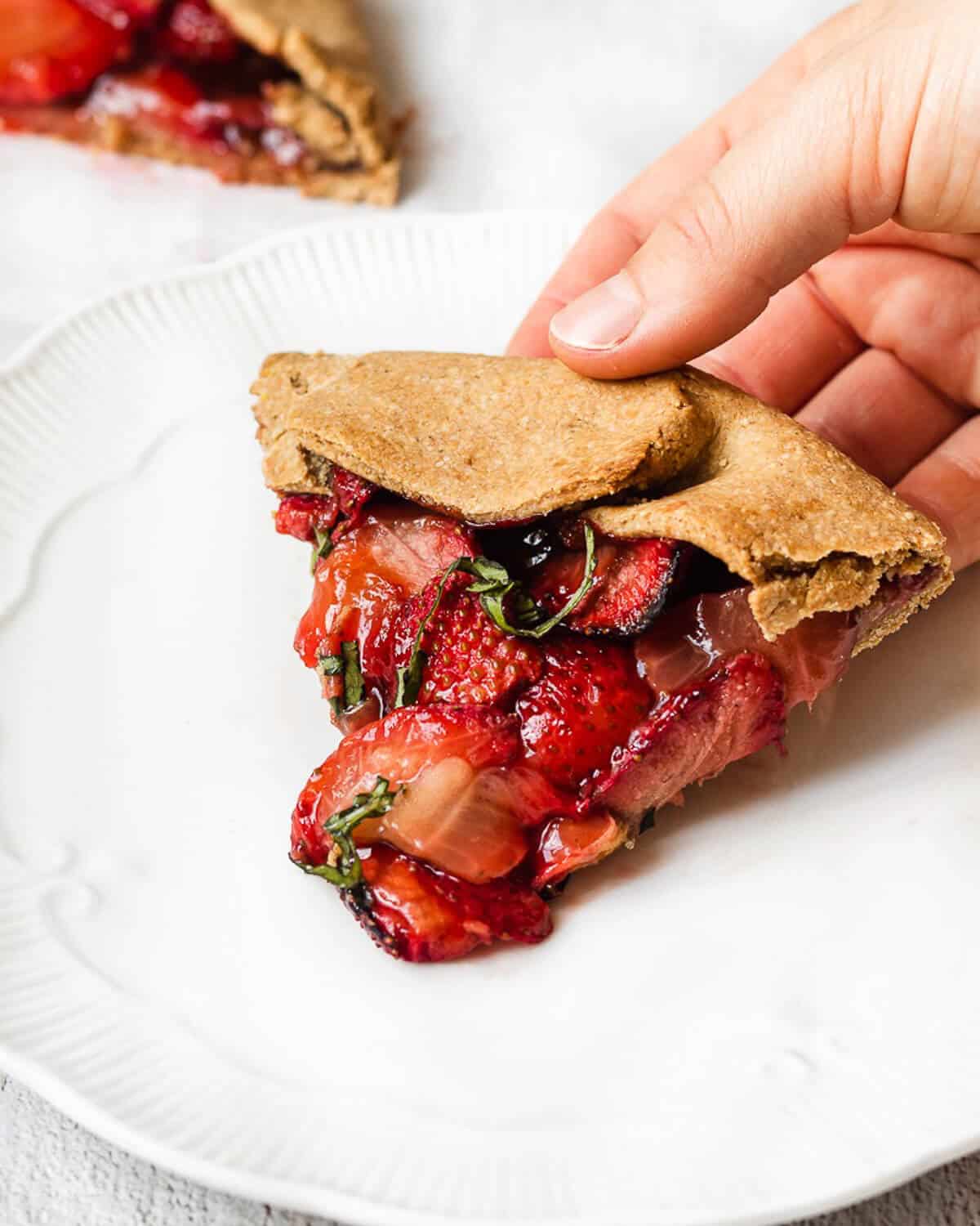 a hand holding a slice of strawberry galette on a white plate