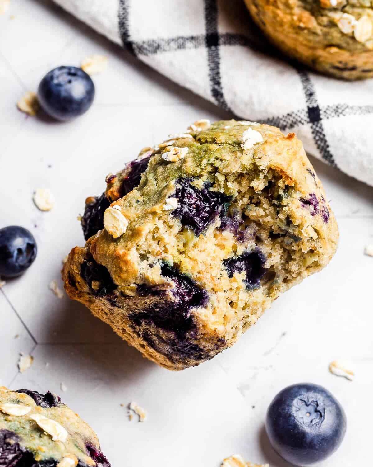 A blueberry banana muffin with a bite missing lying on its side. There is another muffin in the top right hand corner and another in the bottom left corner. There are blueberries and oats scattered around.