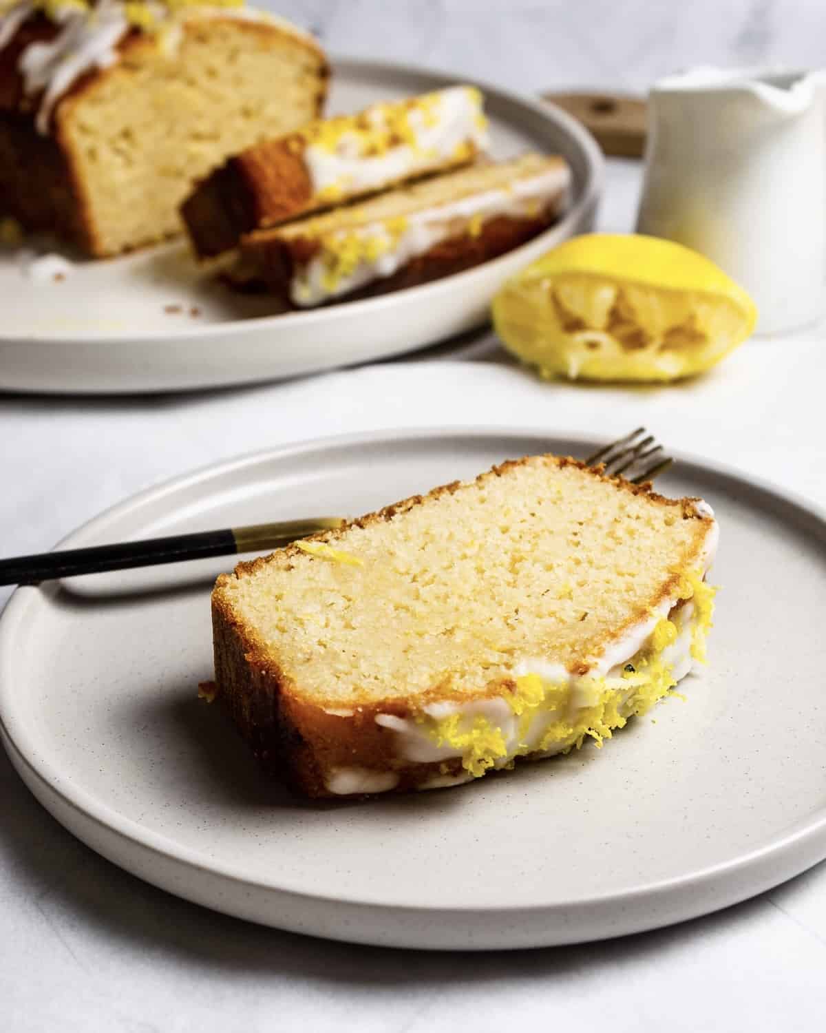 A slice of gluten-free lemon drizzle cake on a plate. There is a fork behind the cake. In the background is the rest of the loaf, sauce jug and half a squeezed lemon.