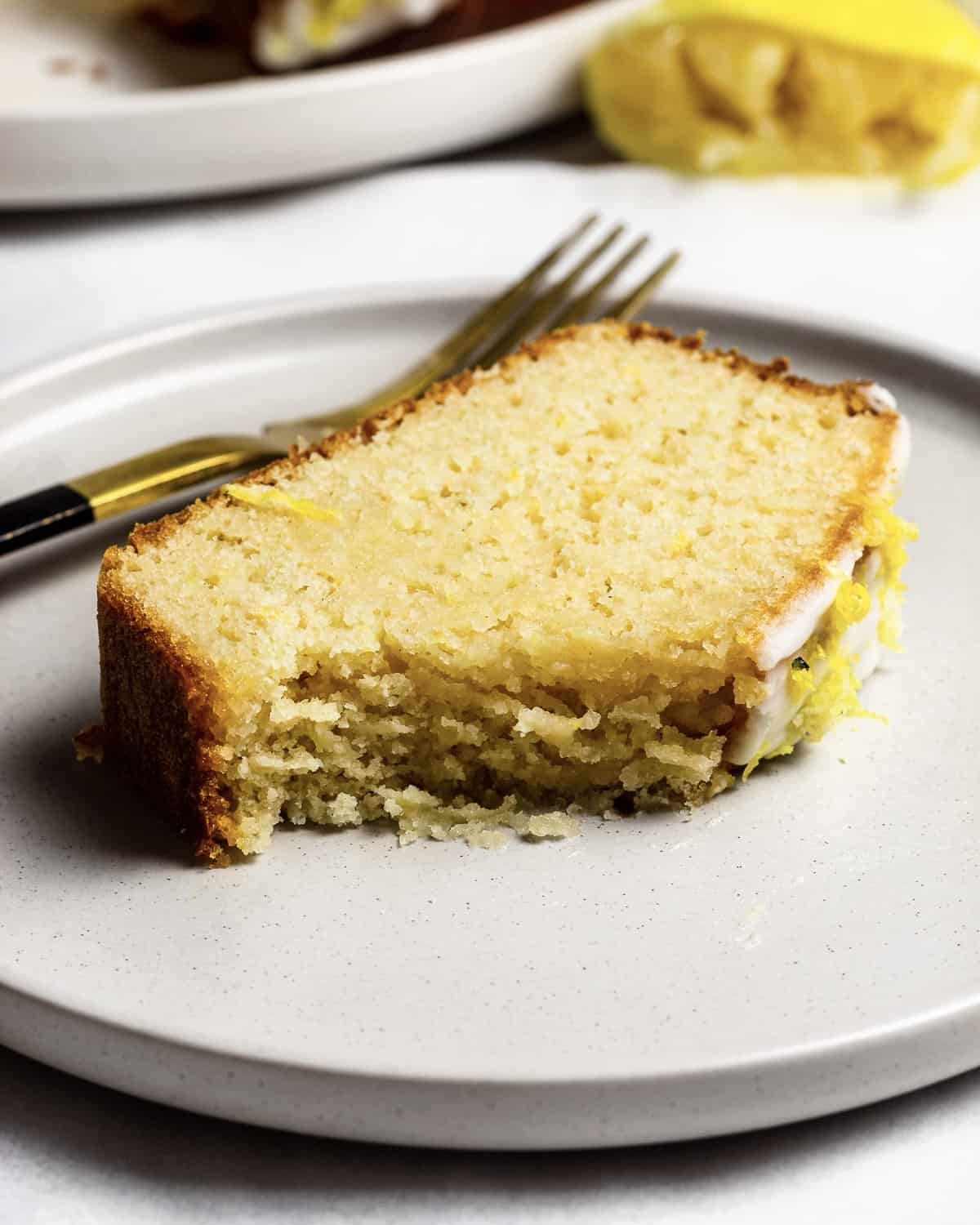 A half eaten slice of fluffy lemon drizzle cake on a plate, there is a fork and lemon in the background.