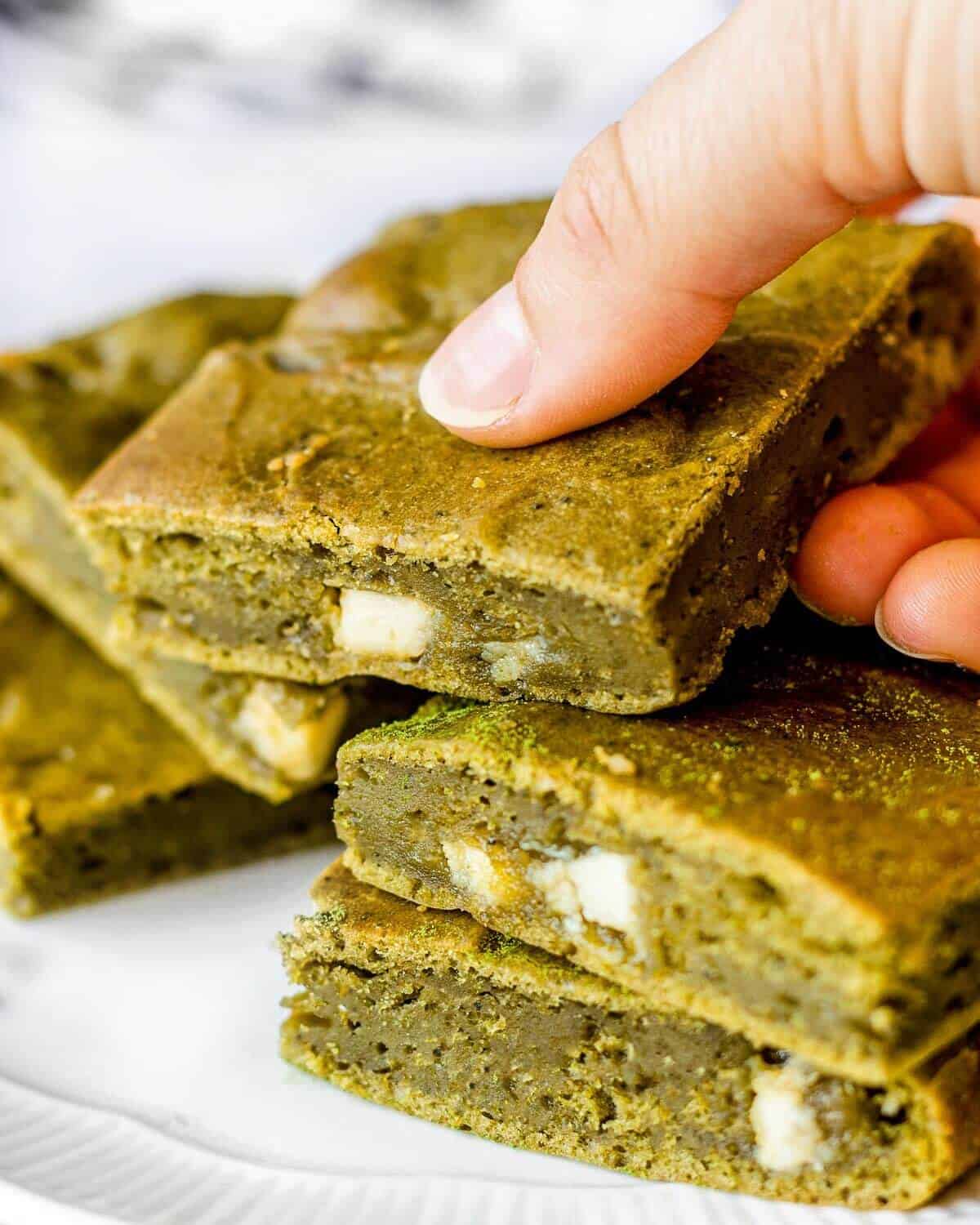 Five matcha brownies with white chocolate stacked on a white plate, someone is holding the one on top.