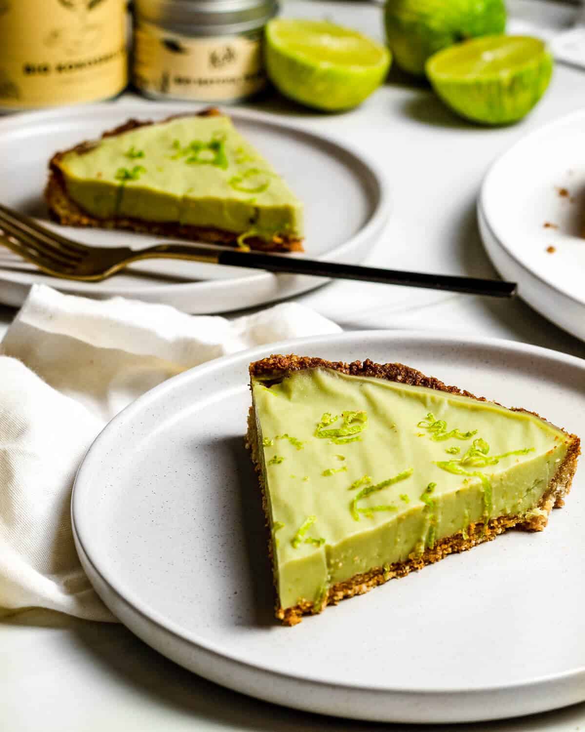 Two plates with a slice of vegan key lime pie on each. There is a fork on the plate in the top left corner. In the background are some limes, coconut milk and matcha.