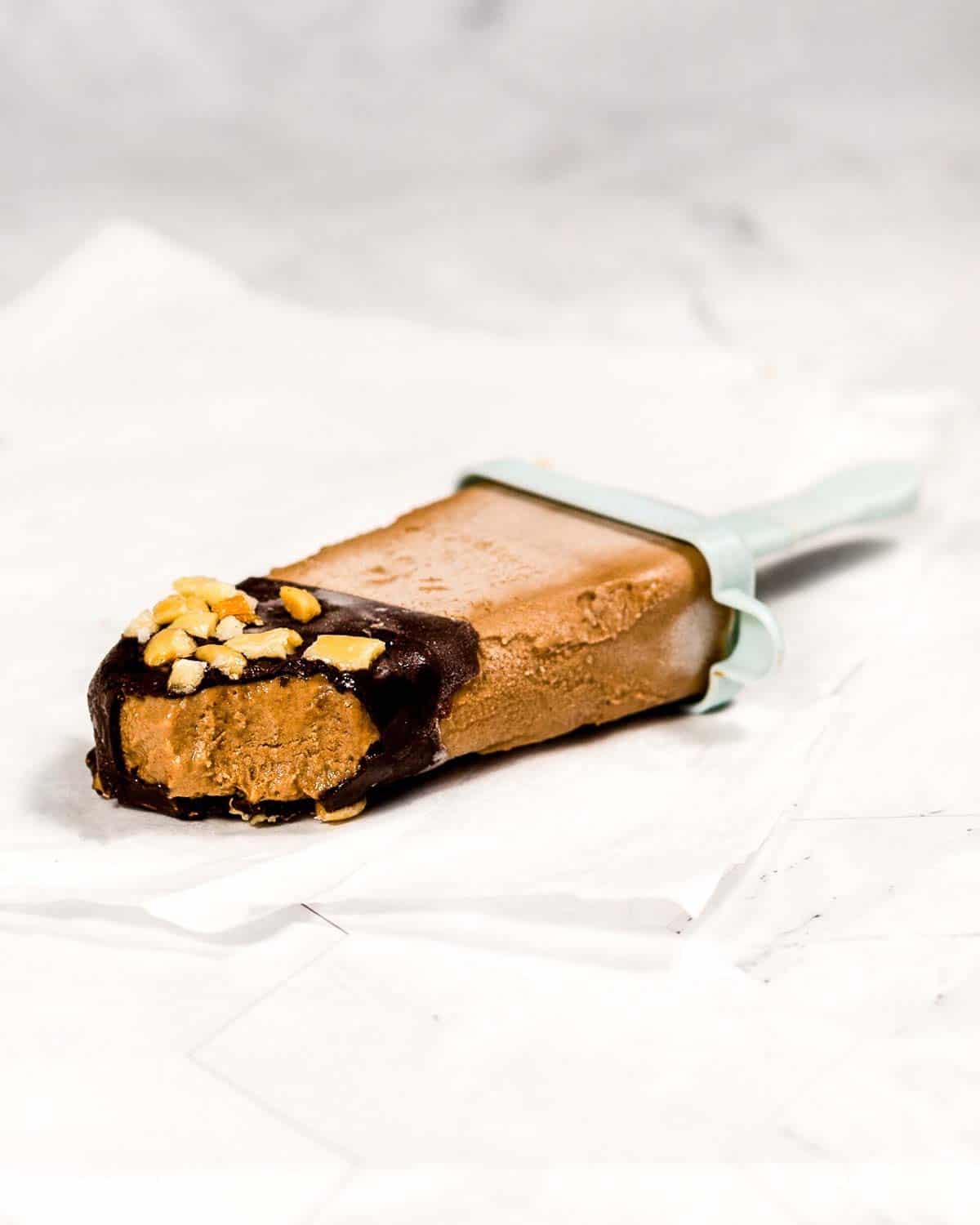 A chocolate ice cream bar dipped in chocolate and peanuts on parchment paper. A bite is missing.