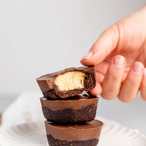 Three peanut butter cup cheesecakes stacked on top of each other on a white plate. A hand is slightly holding the top one and a bite is missing reavealing the cheesecake filling.