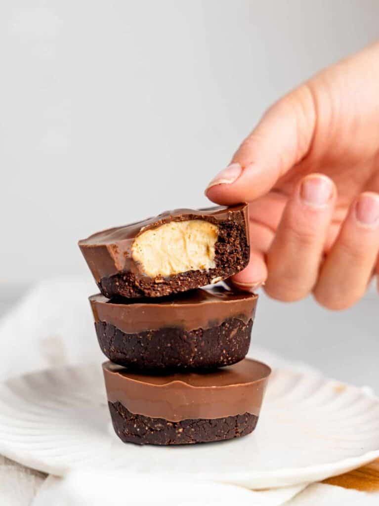 Three peanut butter cup cheesecakes stacked on top of each other on a white plate. A hand is slightly holding the top one and a bite is missing reavealing the cheesecake filling.